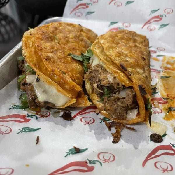 Two crispy birria tacos with melted cheese and shredded meat on chili-patterned paper at Casa Agave.