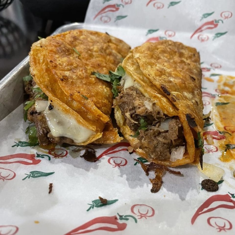 Two crispy birria tacos with melted cheese and shredded meat on chili-patterned paper at Casa Agave.