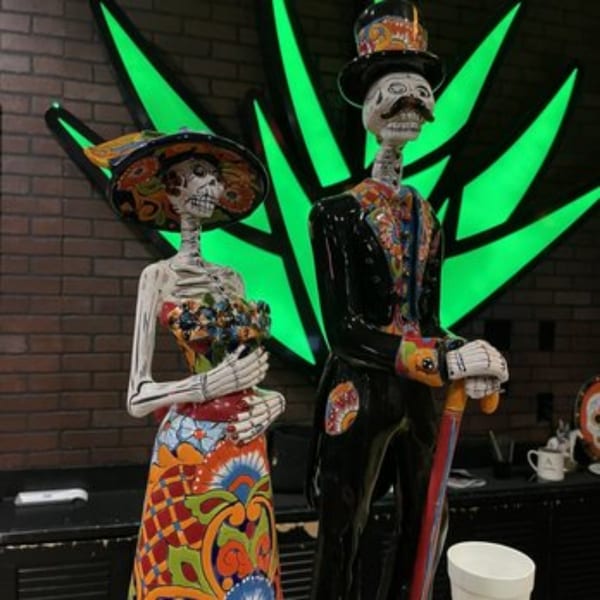 Two colorful skeleton statues, a female and a male, stand in front of a glowing green agave plant.